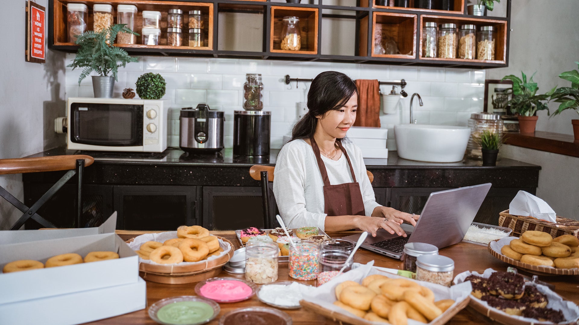 Woman baker sitting in her kitchen and working on her laptop with trays of donuts, sprinkles, and frosting.